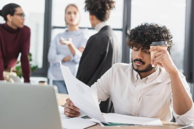 Ofiste dizüstü bilgisayarın yanında kahve ve belge tutan yorgun Hintli işadamı 