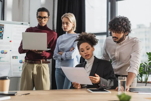 African american colleague holding document near indian colleague and digital tablet in office