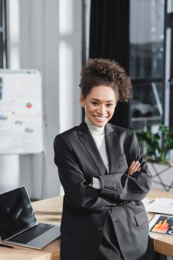 Mutlu Afro-Amerikan iş kadını laptopun yanındaki kameraya ve ofisteki evraklara bakıyor. 