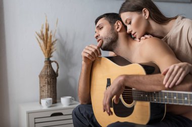 Evde akustik gitar tutan tişörtsüz bir adam var. 