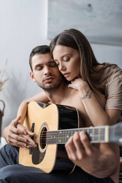  Young woman hugging boyfriend and playing acoustic guitar at home 