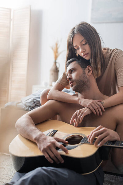 Brunette woman hugging shirtless man holding acoustic guitar in bedroom 