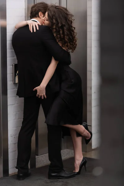 passionate woman in black slip dress lifting leg and touching buttocks of man in suit near elevator