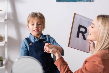 Gülümseyen konuşma terapisti danışma odasındaki aynanın yanında çocuğun elini ve panosunu tutuyor. 