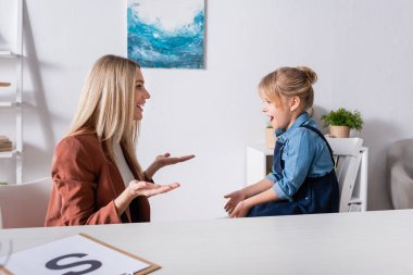 Side view of cheerful speech therapist talking with girl in consulting room  clipart