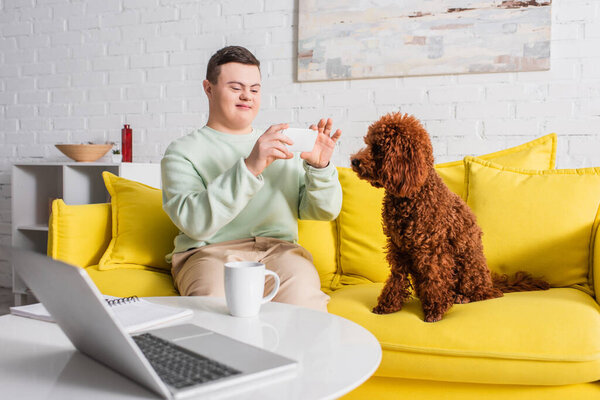 Teenager with down syndrome taking photo of poodle near laptop and cup at home 