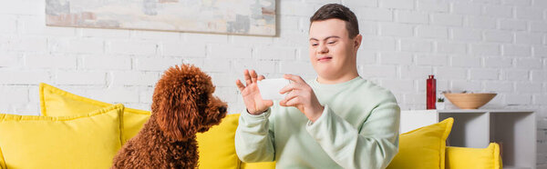 Smiling teenager with down syndrome taking photo of poodle at home, banner 