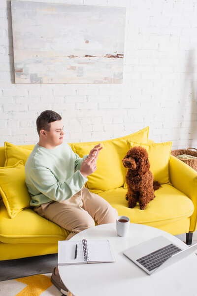 Teenager with down syndrome taking photo of poodle near tea and laptop on table  