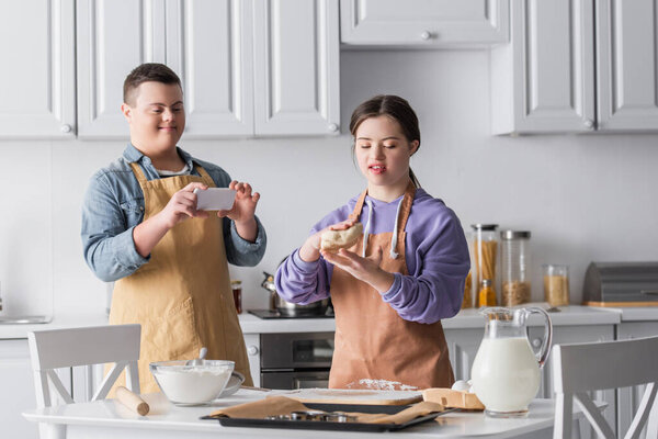 Teen girl with down syndrome holding dough near friend with cellphone in kitchen 