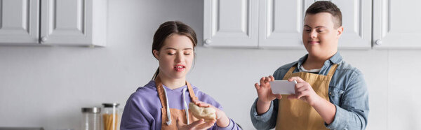 Positive girl with down syndrome holding dough near friend with mobile phone in kitchen, banner 