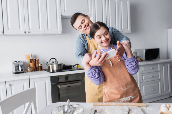 Cheerful couple with down syndrome taking selfie on smartphone near dough in kitchen 