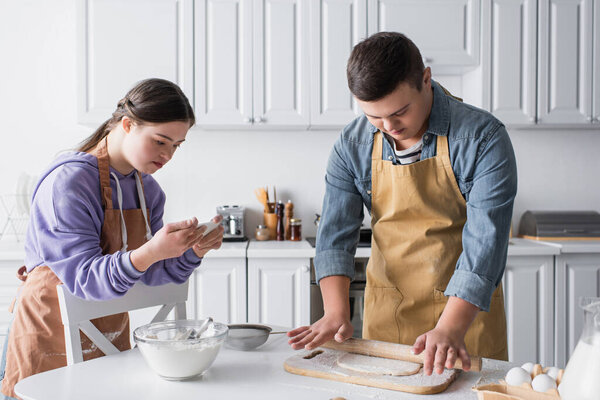 Teenager with down syndrome using smartphone near friend cooking dough in kitchen 