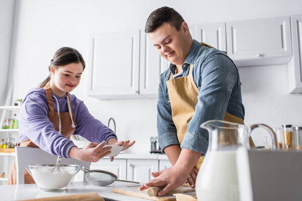 Smiling teenager with down syndrome rolling dough near friend taking photo in kitchen 
