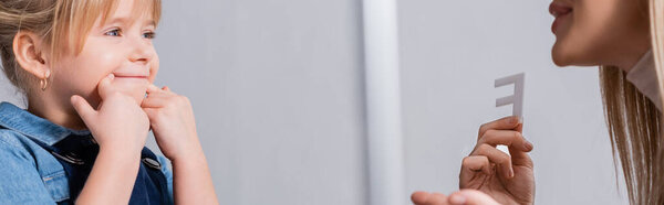 Речевой терапевт держит письмо рядом с ребенком касаясь рта в консультационной комнате, баннер 