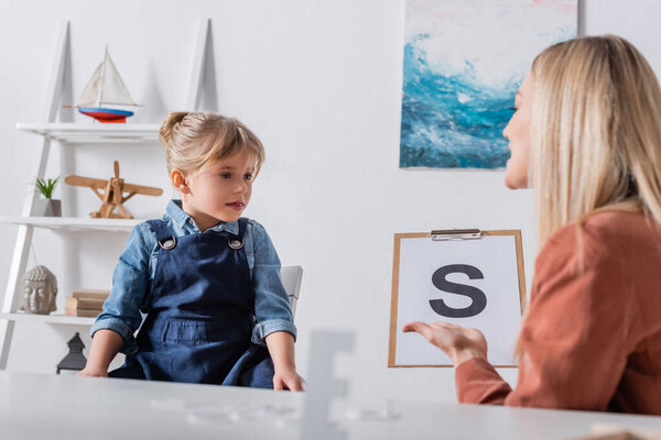 Girl sitting near blurred speech therapist with letter on clipboard in consulting room 