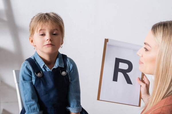 Speech therapist holding clipboard with letter near girl in consulting room 