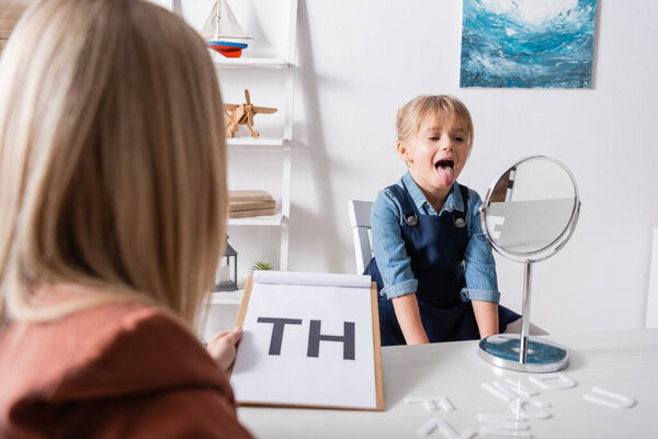 Pupil sticking out tongue near mirror and blurred speech therapist in classroom 