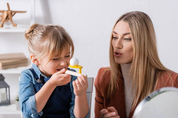Smiling kid using respiratory muscle trainer near logopedist in classroom 
