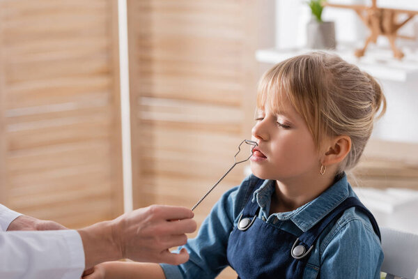 Speech therapist in white coat holding tool near kid in classroom 