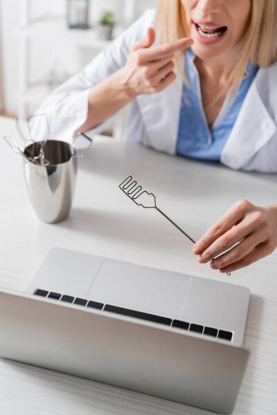 Cropped view of blurred speech therapist holding logopedic probe and pointing at mouth during video call on laptop 