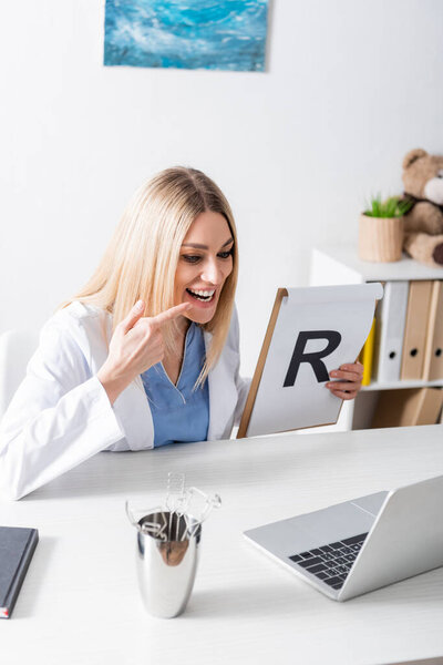 Speech therapist holding clipboard with letter and pointing at mouth near laptop and tools in consulting room 