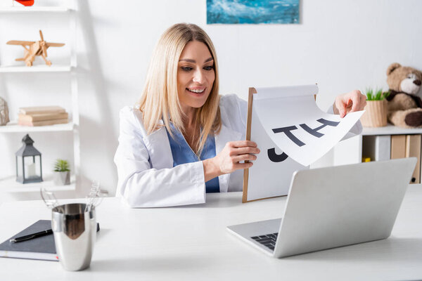 Cheerful logopedist holding clipboard with letters during video call on laptop in consulting room 