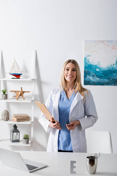 Cheerful speech therapist holding clipboard near laptop on table in consulting room 