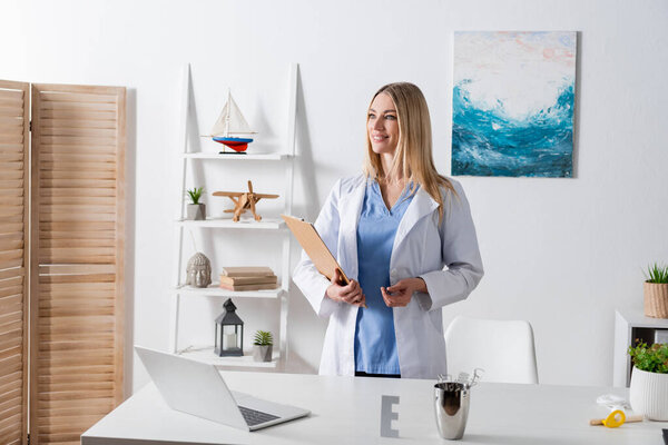 Cheerful logopedist holding clipboard near tools and laptop in consulting room 