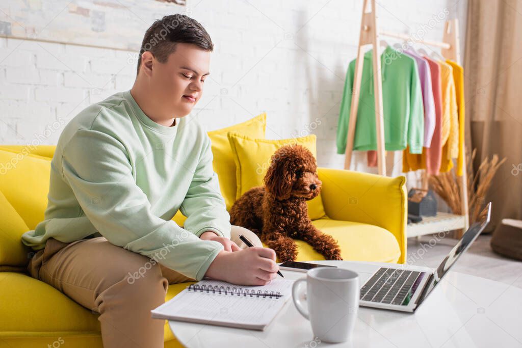Teenage boy with down syndrome writing on notebook near poodle and devices at home 