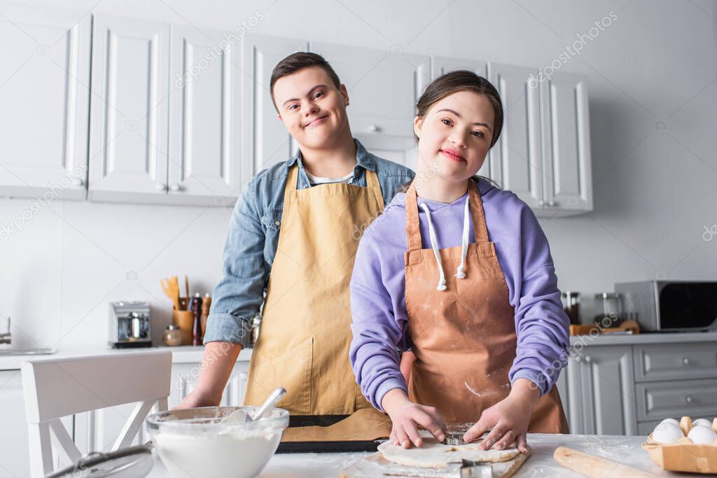 Cheerful couple with down syndrome looking at camera while cooking in kitchen 