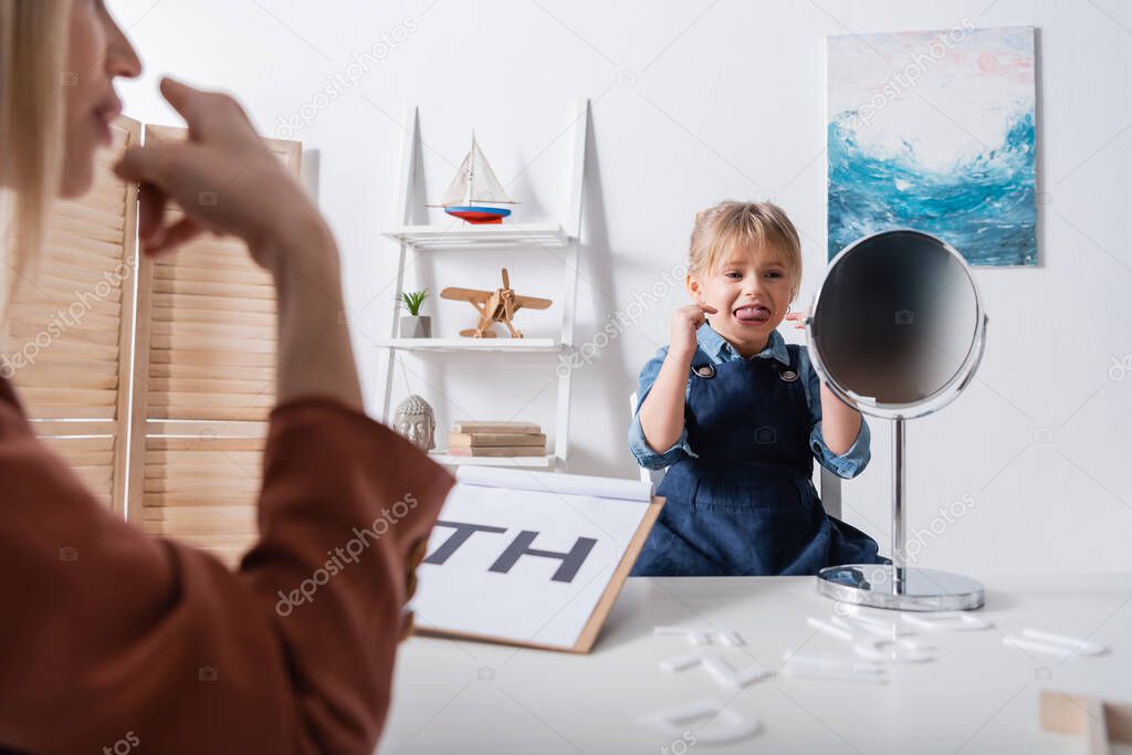 Pupil sticking out tongue and pointing with fingers while talking near mirror and blurred speech therapist in classroom 