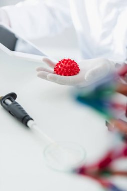 red coronavirus bacteria model in hand of cropped virologist on blurred background clipart