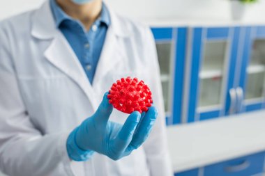 Kırmızı koronavirüs bakteri modeli tutan mavi lateks eldivenli bulanık biyomühendisin kısmi görüntüsü