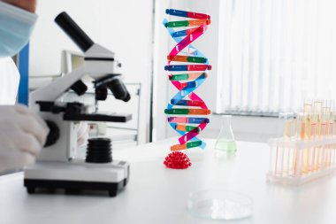 Tıbbi maskeli bilim adamının DNA ve koronavirüs bakteri modelleri yakınında mikroskopla çalışırken görüntülenmiş görüntüsü.