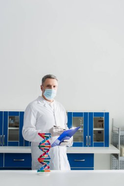 Tıbbi maskede biyomühendis. Pano ve kalemi biyoloji laboratuvarında tutuyor. 