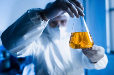 Tehlikeli madde giysileri içinde bulanık biyoteknoloji uzmanı elinde portakal suyu şişesi tutuyor.