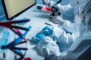Tehlikeli madde giysileri içindeki biyo-mühendisler laboratuvarda gri ekranlı bilgisayar monitörünün yanında çalışıyorlar.