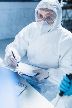 Tehlikeli madde giysisi içindeki genetikçi elinde boş bir not tahtası ve kalemle laboratuvarda bekliyor.