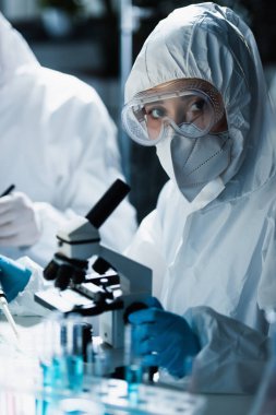 Tehlikeli madde giysisi, gözlük ve tıbbi maske takan kadın mikroskobun yanındaki kameraya bakıyor.