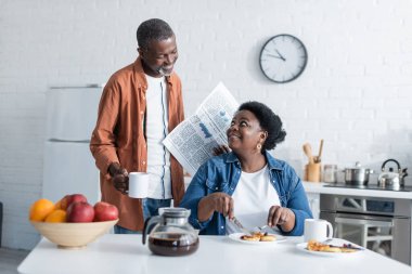 Neşeli ve kıdemli Afro-Amerikalı adam elinde fincanla gazete tutarken karısının kahvaltısını seyrediyor. 