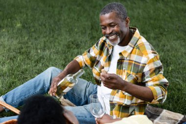 Mutlu kıdemli Afrikalı Amerikalı adam karısının yanında bir şişe şarap tutuyor.