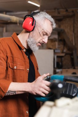 carpenter in protective earmuffs working with miter saw in workshop clipart