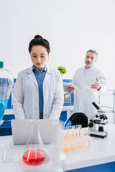 blurred bioengineer talking to colleague using laptop near microscope and test tubes