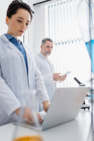 young scientist using laptop near mature colleague with digital tablet on blurred foreground