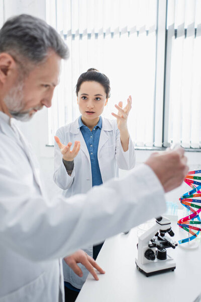 young geneticist gesturing while talking to mature colleague on blurred foreground