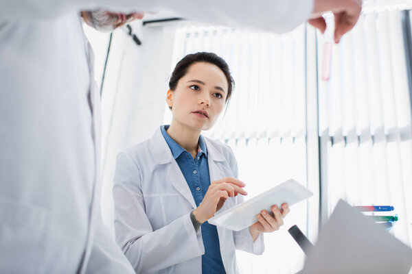 bioengineer holding digital tablet near blurred colleague with test tube