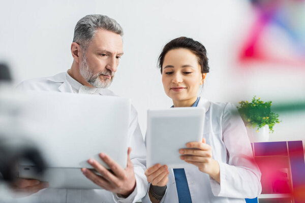 smiling scientist showing digital tablet to mature colleague with laptop