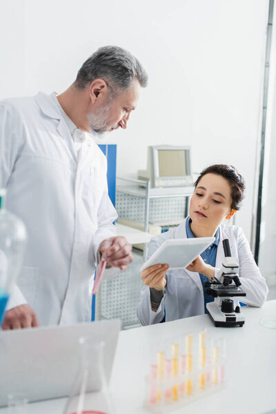 young bioengineer showing digital tablet to mature colleague near microscope