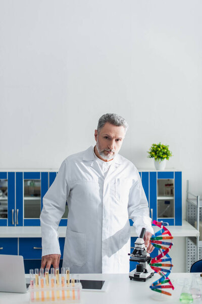 bearded geneticist in white coat looking at camera near dna model and microscope