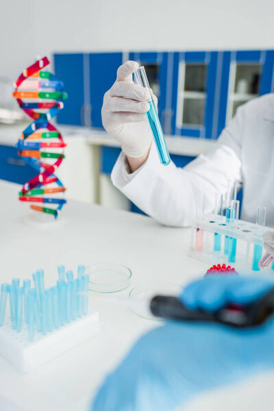 partial view of scientist holding test tube near dna model on blurred foreground